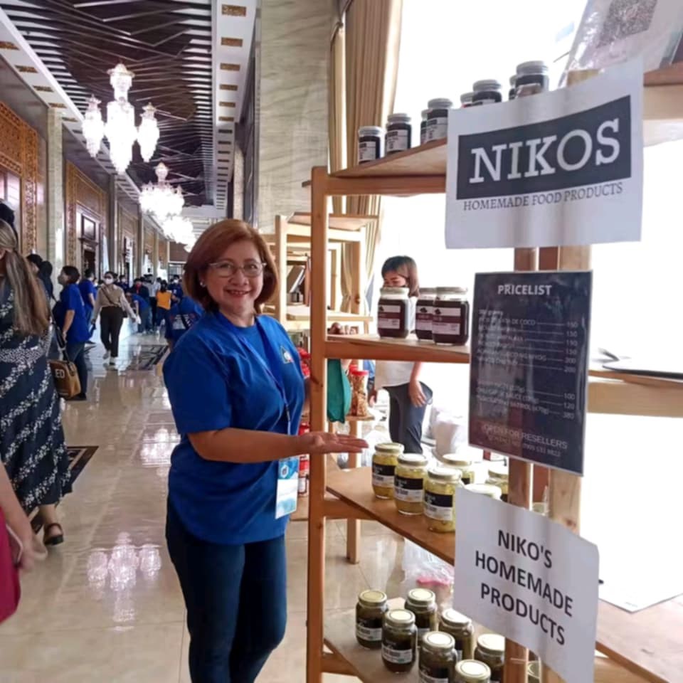 A smiling woman stands gesturing towards a wooden display shelf showcasing "NIKOS HOMEMADE FOOD PRODUCTS". She is wearing glasses, a blue shirt, and dark pants. The shelves are stocked with jars of various food items, and a pricelist is visible on the side of the shelf. A sign clearly reads "NIKOS HOMEMADE FOOD PRODUCTS". The background shows a well-lit indoor space with ornate architectural details, including patterned ceilings and chandeliers, and several people walking in the distance. The setting appears to be a trade show or a market within a formal building. Given the previous images and context, the woman is likely Filipina and the products are homemade food items from "Nikos".