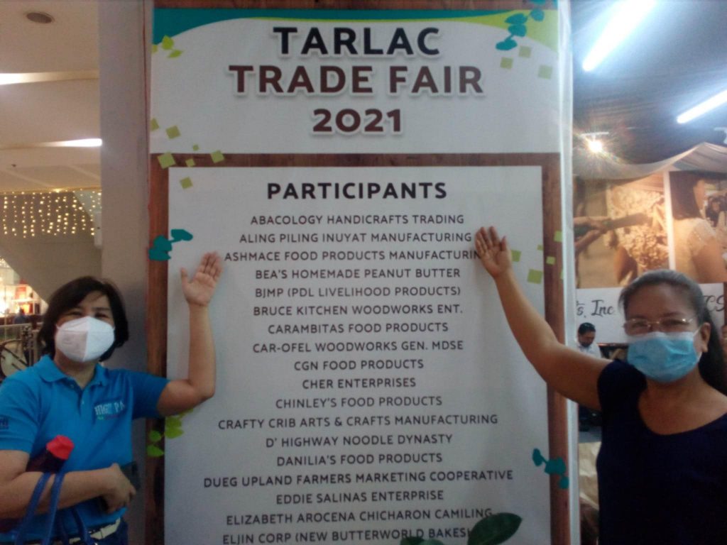 Two individuals, likely women, wearing face masks, stand on either side of a sign that reads "TARLAC TRADE FAIR 2021". The sign lists numerous participants, including various food product manufacturers, handicraft businesses, and other enterprises. The women are smiling behind their masks and have one hand raised in a welcoming gesture towards the sign. The setting appears to be a brightly lit indoor space, possibly a mall or exhibition center, given the pillars and lighting fixtures visible in the background. The sign indicates a trade fair event that took place in Tarlac, likely in the Philippines. Based on the visual cues and the likely location, the individuals are likely Filipina.