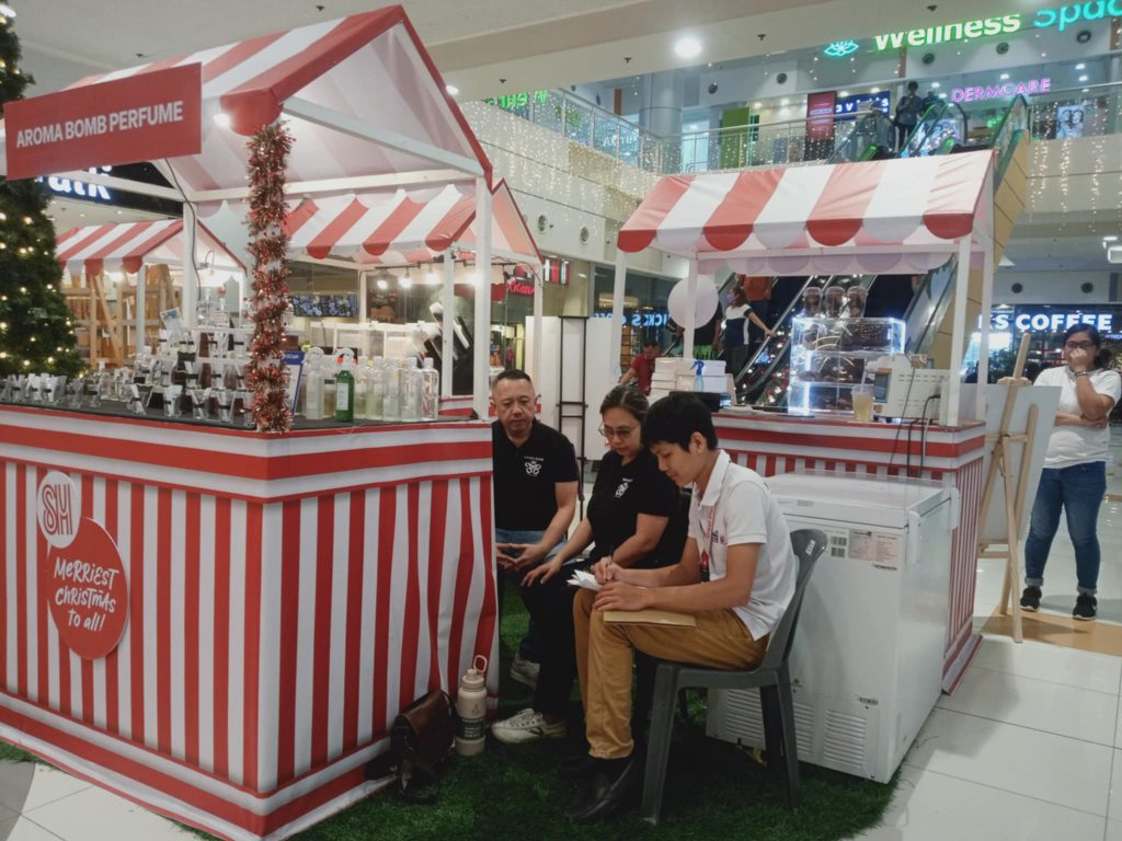 Three people at "AROMA BOMB PERFUME" kiosk in a mall. One seated writing, two standing looking at paper.