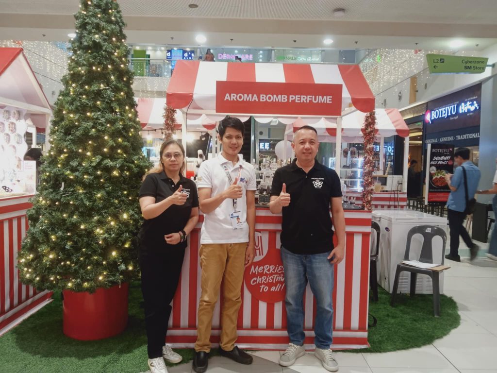 Three people at "AROMA BOMB PERFUME" kiosk in a mall, giving thumbs up. Christmas tree on the left.