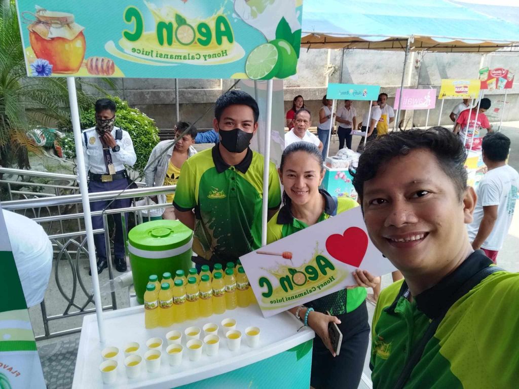 A selfie captures three smiling individuals at an outdoor booth for "Agon C Honey Calamansi Juice," with samples and promotional materials visible.