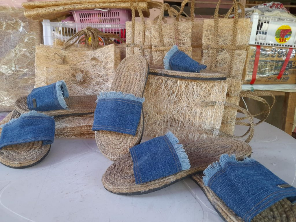 Several pairs of handmade slippers with jute soles and denim straps are displayed on a light-colored surface. The slippers have a rustic, artisanal appearance, with frayed edges on the denim straps. Behind the slippers, various woven bags and other handcrafted items made from natural fibers are visible, suggesting a collection of eco-friendly or traditionally made products. The background is somewhat cluttered with more of these woven materials. The lighting is bright and even, focusing attention on the slippers and bags. Given the context of the previous images and the likely location, these could be Filipino-made crafts.