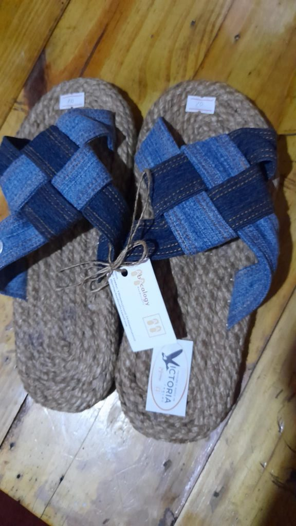 A pair of handmade slippers rests on a wooden surface. The slippers have thick soles made of tightly wound jute or a similar natural fiber. The straps are crafted from woven blue denim fabric, creating a crisscross pattern over the top of the foot. A white tag with black text is attached to one of the slippers with a piece of string. The slippers appear to be artisanal and eco-friendly. Given the likely origin based on the previous images, these could be Filipino-made.