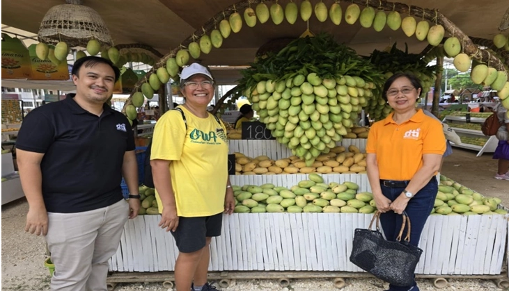 Three individuals stand in front of a display booth at the Manggahan Festival 2025, promoting Guimaras mangoes. The booth features an arch made of hanging mangoes and a table filled with neatly arranged mangoes.