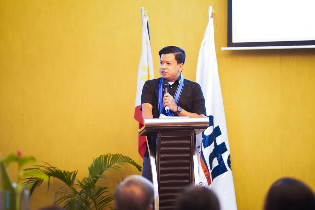 DTI Assistant Secretary Kristian R. Ablan delivers a speech at the podium during the Palawan Investors' Roadshow, with Philippine flags in the background and an audience in the foreground, highlighting the culmination of the energy efficiency project for MSMEs.