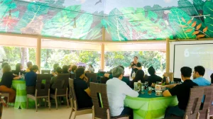 A group of people attending a presentation at Daluyon Resort, seated around tables covered with green tablecloths. The venue is an open-air pavilion with a colorful ceiling featuring large leaf patterns. A speaker stands in front of a projector screen displaying the number 6 and the word 'hazardous'.