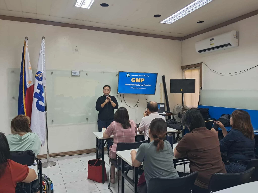 A presenter delivers a seminar on "GMP Good Manufacturing Practice" to MSMEs in a classroom setting, with participants seated at desks and a presentation slide displayed on a screen. Two flags are positioned near a whiteboard behind the speaker.
