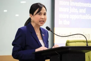 DTI Secretary Cristina A. Roque speaking at the podium during the CREATE MORE High-Level Forum on March 28. She is wearing a navy blue blazer and standing in front of a microphone, with a presentation screen in the background displaying text about investments, jobs, and opportunities.