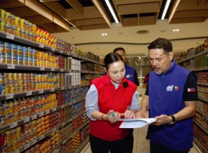 DTI Secretary Cristina A. Roque and Assistant Secretary Agaton Teodoro O. Uvero conducting price and supply monitoring in a grocery store aisle in Muntinlupa City on May 29. Secretary Roque, in a red vest, examines a canned product, while Assistant Secretary Uvero, in a blue vest with a Philippine flag patch, holds a clipboard. Shelves stocked with canned goods are visible in the background.