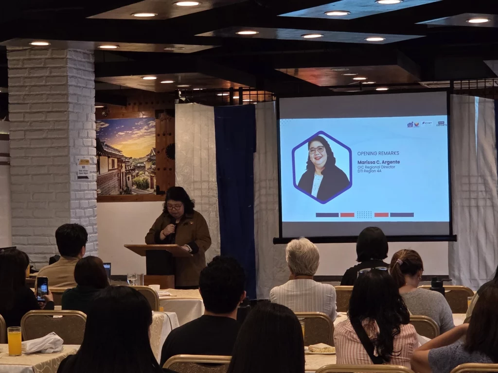 DTI Region 4A Director Marissa Argente delivers opening remarks during the FTA Integrated Platform Roadshow at Hotel Marciano, addressing an audience of exporters and trade stakeholders in a conference room with a presentation screen displaying her name.