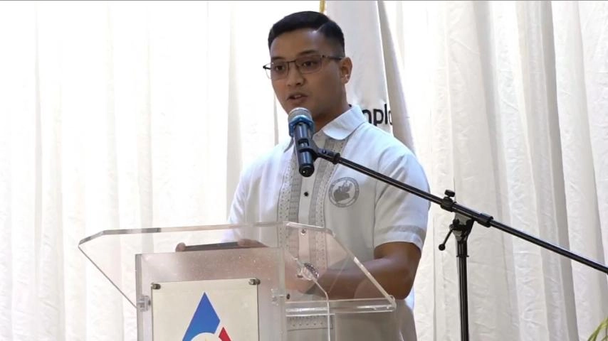 A man wearing glasses, a white collared shirt with a logo, and a microphone is speaking at a clear podium with a logo visible at the bottom. He is standing in front of a white curtain with a partially visible flag or banner to his right.