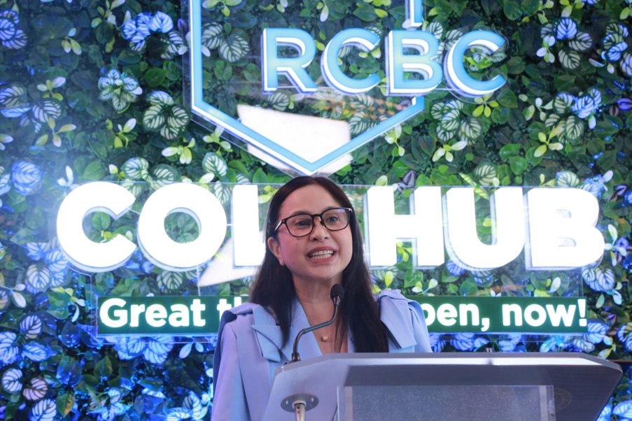 Department of Trade and Industry (DTI) Secretary Cristina A. Roque speaking at a podium during the launch event of the RCBC x BahAI Real Estate Financing and Property-Matching Platform. The backdrop features the RCBC logo and greenery with the words "COLLAB HUB" and "Great things happen now!"