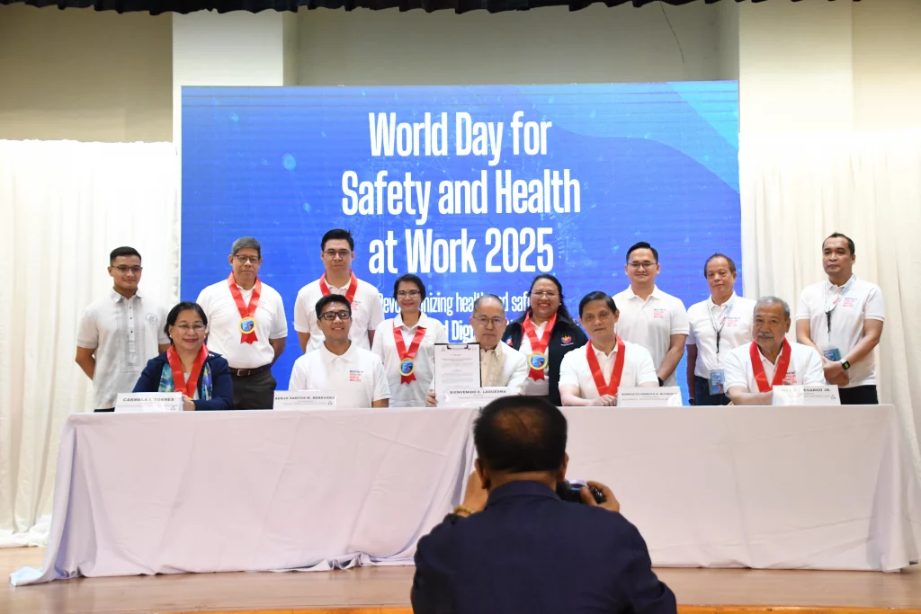 A group of people are standing and sitting behind a long table covered with a white cloth. Behind them is a large blue banner that reads "World Day for Safety and Health at Work 2025" and includes the theme "Revolutionizing Health and Safety: The Role of Artificial Intelligence (AI) and Digitalization at Work Towards Achieving Zero Accident." Most individuals are wearing white shirts and red lanyards with IDs. Some individuals in the front row are holding documents. A person in the foreground, with their back to the camera, appears to be taking a photo of the group with a camera. The setting suggests a formal event or press conference related to workplace safety and health.