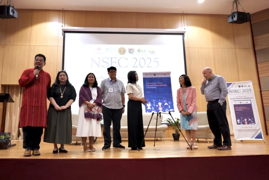 A group of individuals are standing on a stage in front of a large screen displaying "NSEC 2025" and the theme "Social Enterprises: Scaling Change in the Philippines." The people on stage appear to be a diverse group, possibly speakers or organizers of the event. One man on the far left is speaking into a microphone.