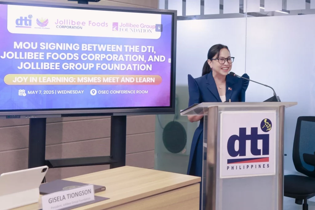 Secretary Cristina A. Roque delivering a speech at a podium during the MOU signing event between the Department of Trade and Industry (DTI), Jollibee Foods Corporation (JFC), and the Jollibee Group Foundation (JGF). The backdrop displays the event details and logos of the involved organizations.