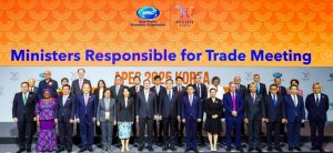 A group photo of delegates at the "Ministers Responsible for Trade Meeting" held by the Asia-Pacific Economic Cooperation (APEC) in 2023. The backdrop features an orange gradient with circular patterns and the text "APEC 2023 KOREA." The delegates are standing in two rows, dressed in formal attire, with some wearing traditional clothing.