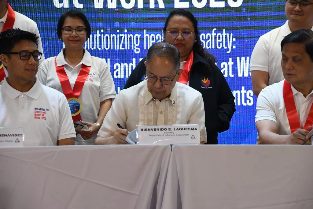 A close-up shot captures Bienvenido E. Laguesma, identified by a nameplate as the Secretary of Labor and Employment, signing a document at a table covered with a white cloth. He is wearing glasses and a light-colored embroidered shirt. Several other individuals are partially visible standing behind the table, observing the signing. A large blue banner in the background displays "World Day for Safety and Health at Work 2025." The focus is on the signing of the document, suggesting a formal agreement or declaration related to the event.