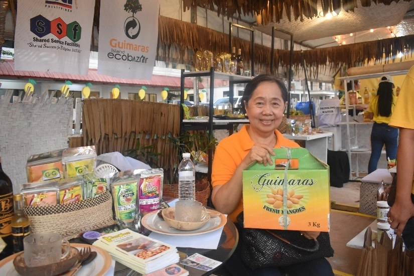 DTI Region 6 Director Rachel N. Nufable showcases a box of premium Guimaras mangoes at the DTI booth, surrounded by promotional materials and local products during the Manggahan Festival 2025.