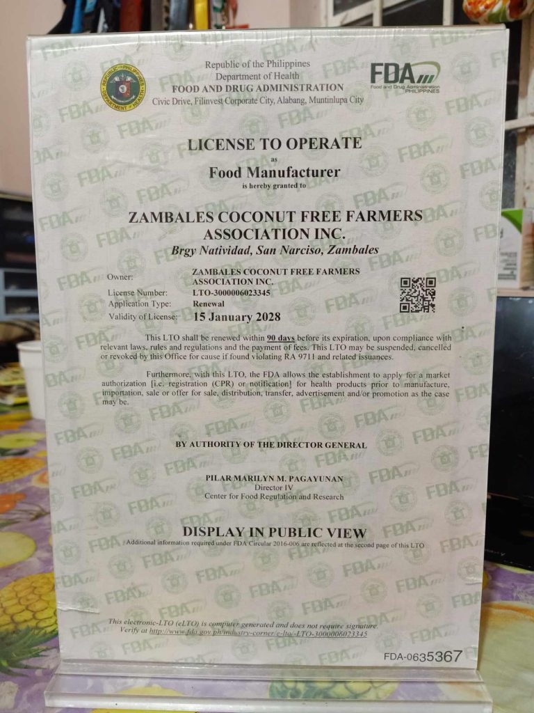 A clear photo of a "License to Operate" certificate issued by the Food and Drug Administration (FDA) of the Republic of the Philippines to "ZAMBALES COCONUT FREE FARMERS ASSOCIATION INC." for food manufacturing. The certificate displays the FDA logo and official seals, the name and address of the licensee in Brgy Natividad, San Narciso, Zambales, the license number "LTO-3000006893342," the application type "Renewal," and the validity date "15 January 2028." A QR code is also visible on the right side of the document. The document is displayed in a clear plastic stand.