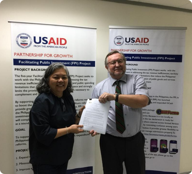 Two people smiling and shaking hands while holding a document in front of USAID 'Partnership for Growth' project banners. Two people smiling and shaking hands while holding a document in front of USAID 'Partnership for Growth' project banners.