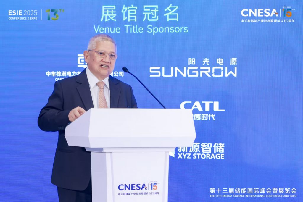 A medium shot shows Glenn G. Peñaranda, who appears to be of East Asian descent, at a podium with a microphone. He is wearing a suit and tie. The background features a blue screen with text in English and Chinese, including "ESIE 2025" and "THE 13TH ENERGY STORAGE INTERNATIONAL CONFERENCE AND EXPO," along with sponsor names. The setting is a formal conference or presentation.