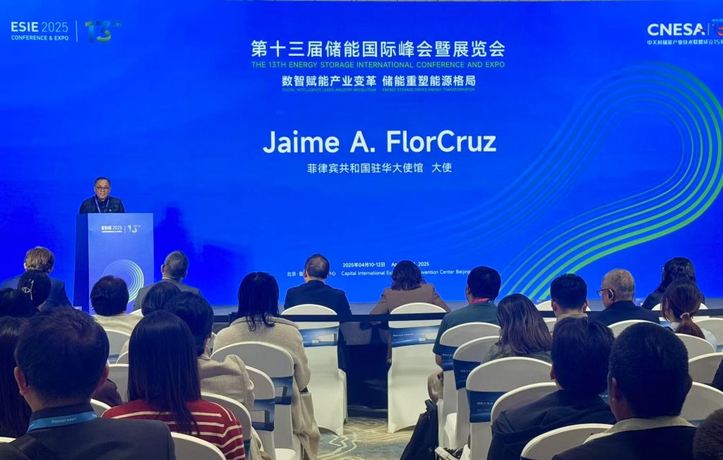 A medium shot of a conference hall with an audience facing a stage. The stage has a large blue screen displaying text in both English and Chinese: "THE 13TH ENERGY STORAGE INTERNATIONAL CONFERENCE AND EXPO" and "Jaime A. FlorCruz" with additional text below it.  A man is visible on the left, standing behind a podium and speaking. The audience is seated in rows of chairs, with their backs to the camera. The setting suggests a formal presentation or lecture at an international conference.