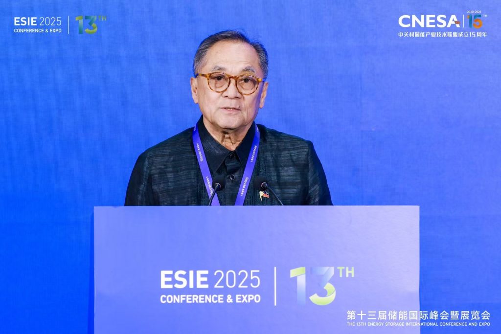 A medium shot captures Jaime A. FlorCruz, who appears to be of East Asian descent, speaking at the "ESIE 2025 13th Conference & Expo". He is positioned behind a light blue podium that displays the conference name and year prominently. FlorCruz is wearing glasses, a black collared shirt, and a lanyard around his neck. Behind him, a large blue backdrop features the conference logo and name in both English and what appears to be Chinese characters, along with "CNESA" and "THE 13TH ENERGY STORAGE INTERNATIONAL CONFERENCE AND EXPO". The lighting is bright, focusing on the speaker.