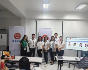 Group photo of DTI Rizal representatives and partners from Meralco, Bayad, and MPioneer Insurance during the Power Coaching session in Morong, Rizal, with participants holding a certificate in front of banners and a presentation slide titled 'Empowering your Business'.
