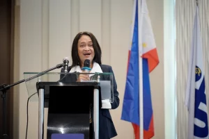 DTI Undersecretary Blesila Lantayona delivers a speech at the 'Mainstreaming Energy Efficiency in MSMEs' Investment Roadshow in Cagayan de Oro, with the Philippine flag and another organizational flag displayed behind the podium.