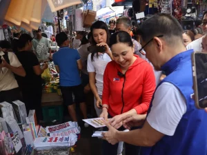 DTI Secretary Cristina A. Roque in a red jacket conducts a price inspection at Divisoria Market, engaging with a vendor while holding the 'Gabay sa Pamimili ng School Supplies' pamphlet, surrounded by school supply stalls and shoppers.