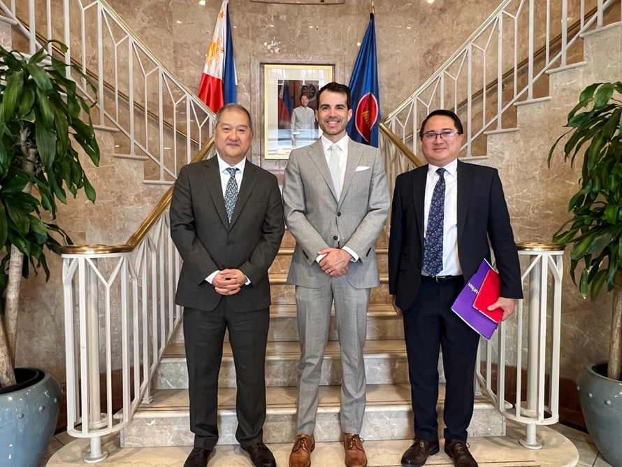 Three men in formal attire standing on a marble staircase with Philippine and ASEAN flags behind them; from left to right: PTIC-Washington D.C. Commercial Counselor Kenneth Yap, Asurion Senior Assistant General Counsel Grant Mullins, and Minister and Consul Hans Siriban.