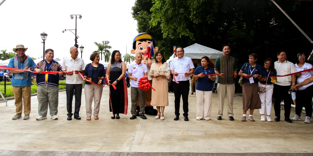 DTI officials and participants, including a mascot, hold a red ribbon during the opening ceremony of the Pampamahalaang Programa at Serbisyo 2025 at Rizal Park, Manila. The outdoor event marks the celebration of Philippine independence and showcases support for MSMEs and the Tatak Pinoy initiative.