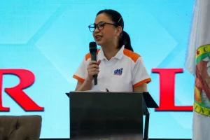 DTI Secretary Cristina A. Roque, wearing a white and orange polo shirt with the DTI logo, speaks into a microphone at a podium during the 'Lab For All' event in Las Piñas. The background displays large red letters spelling 'LAB FOR ALL' and part of a flag is visible on the right.