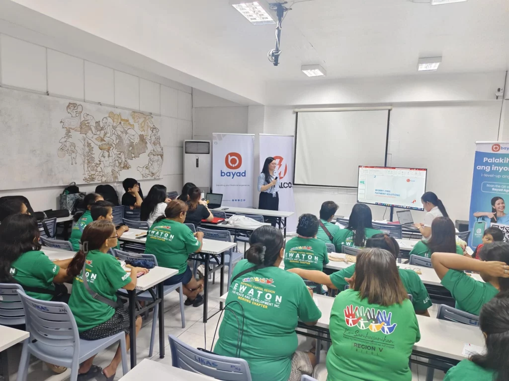 A classroom filled with entrepreneurs wearing green shirts, seated at desks and facing a presenter. The presenter stands near a projector screen displaying information. Behind the presenter are banners for Bayad and PLDT Smart Foundation. The room has white walls, a world map on one wall, and fluorescent lighting. This image is related to an article about DTI Rizal and private partners empowering entrepreneurs through training sessions or workshops.