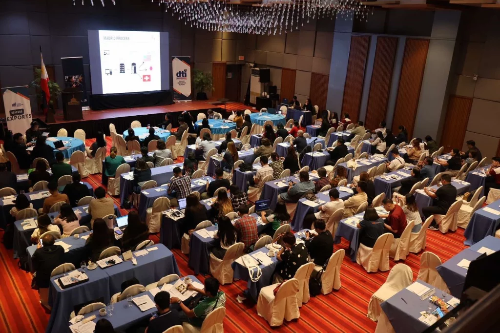 A large conference room filled with attendees seated at tables with blue tablecloths, all facing a stage with a presentation screen. The setting, featuring a red and orange striped carpet and hanging ceiling lights, reflects a DTI-led exporter support event aligned with the Philippine Export Development Plan 2023–2028.