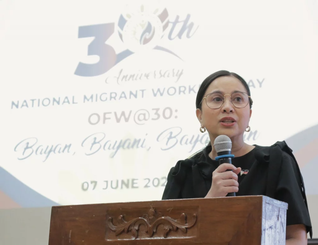 DTI Secretary Cristina A. Roque speaks at a podium during the 30th Anniversary of National Migrant Workers Day, with a screen in the background displaying the theme 'OFW@30: Bayan, Bayani, Bayanihan' and the date '07 June 2023.'