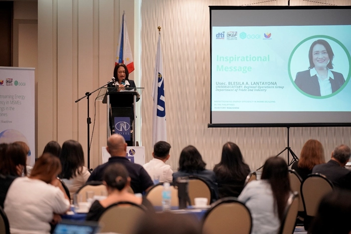 DTI Undersecretary Blesila A. Lantayona delivers an inspirational message during the 'Mainstreaming Energy Efficiency in MSMEs' Investment Roadshow in Cagayan de Oro, with attendees seated at tables and a screen displaying her name and title in the background.