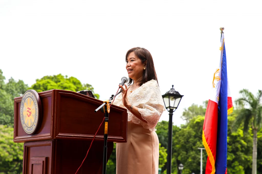 Assistant Secretary Leonila Baluyut of the Department of Trade and Industry delivers a speech at a podium during the opening ceremony of the Pampamahalaang Programa at Serbisyo 2025 in Rizal Park, Manila. The Philippine flag is visible in the background, marking the celebration of the 127th Philippine Independence Day.