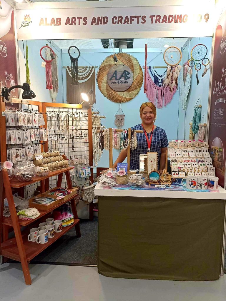 A booth at a craft fair labeled 'ALAB ARTS AND CRAFTS TRADING' displays various handmade items. The booth features dreamcatchers, necklaces, bracelets, earrings, and other jewelry items hanging on racks and displayed on tables. There are also mugs and other small crafts for sale. A person stands behind the table in the center of the booth.