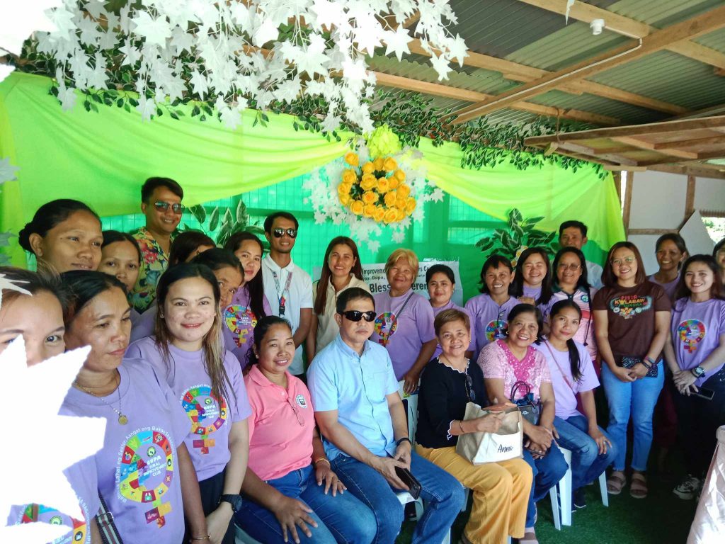 A group of people gathered in a decorated area featuring green and yellow floral arrangements. Some individuals are standing while others are seated, wearing colorful shirts with various designs. The background is adorned with green drapery and hanging white flowers.