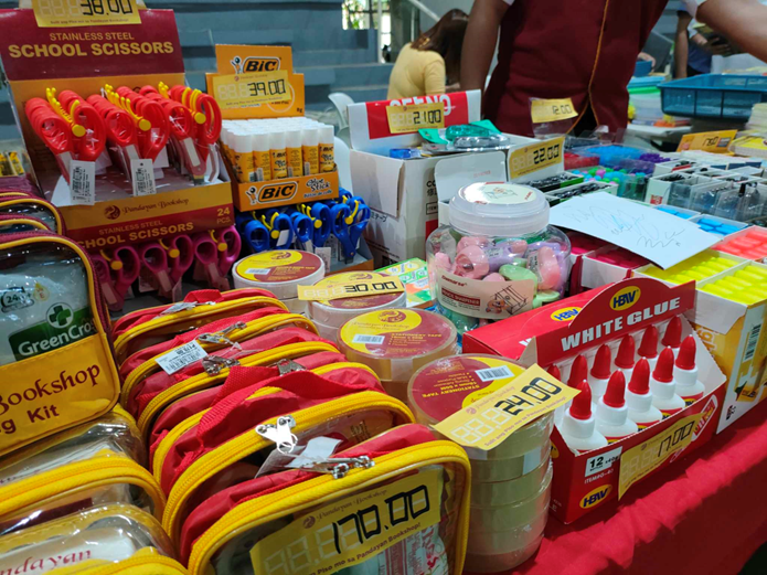 A table filled with various school supplies on display at the DTI Diskwento Caravan. Items include stainless steel scissors, BIC ballpoint pens, glue sticks, correction tape, pencil cases, and white glue. Prices are prominently displayed on yellow tags for each item.