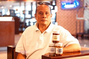 A person in a white polo shirt sits indoors with three jars of Eco Nuestra Homemade Products—labeled "Bagoong" and "Ginamus"—stacked on a table in front. The background features a softly blurred interior with lights and objects.