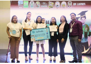 A group of people standing on a stage, holding certificates and a large voucher, with a backdrop showing logos and the text "Our Participants" alongside an illustration of a graduate in cap and gown